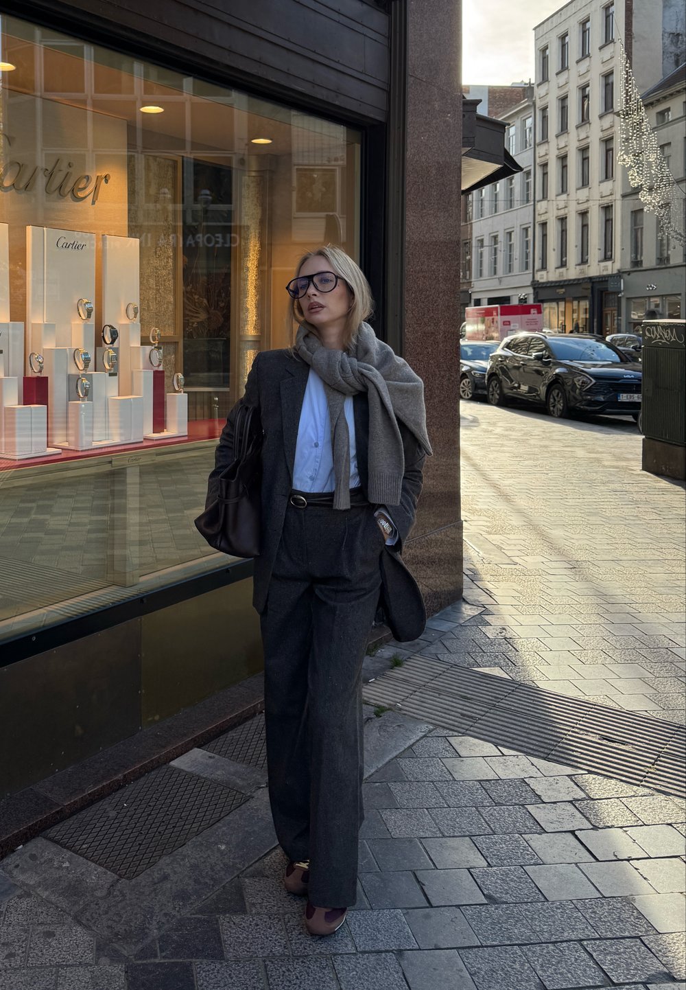 Grå uldblazer og bukser med vide ben, hvid skjorte, lysegråt tørklæde, sort læderhåndtaske og bordeaux loafers, sat op foran en smykkebutik.