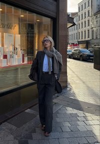 Grå uldblazer og bukser med vide ben, hvid skjorte, lysegråt tørklæde, sort læderhåndtaske og bordeaux loafers, sat op foran en smykkebutik.