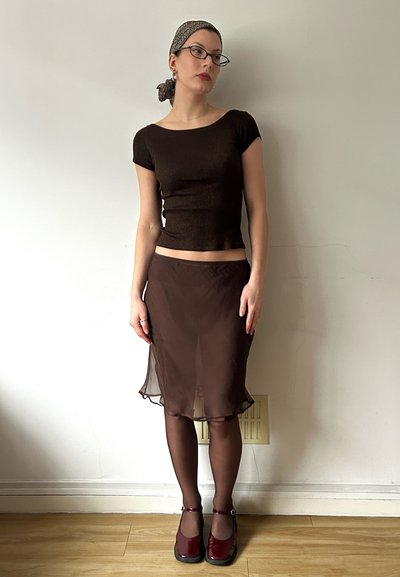 Top corto ajustado marrón de manga corta, falda marrón transparente, medias oscuras marrones y zapatos marón de slip-on. Los accesorios incluyen un pañuelo de cabeza estampado y gafas.