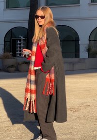 Femme portant des lunettes de soleil, un long manteau sombre, un pull rouge, un pantalon rayé et une écharpe rouge et blanche, tenant une tasse en plein air dans une cour ensoleillée.