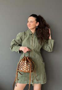 Robe vert olive à fermeture éclair devant avec des manches bouffantes et deux poches latérales, assortie à un sac seau marron orné d'agrafes métalliques.