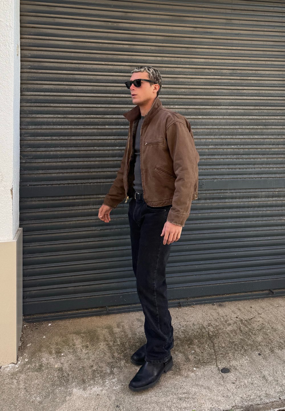 Mann in brauner Jacke, schwarzen Jeans und Stiefeln, der neben einem grauen Metallrollladen steht und dunkle Sonnenbrillen trägt.