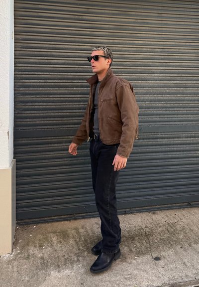 Hombre con chaqueta marrón, vaqueros negros y botas, de pie junto a una persiana metálica gris, llevando gafas de sol oscuras.