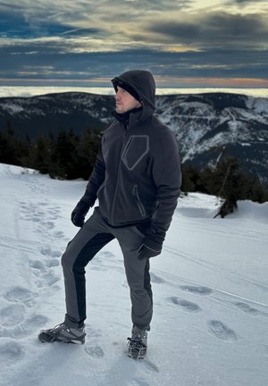 Chaqueta de forro polar negra con capucha, detalles grises, bolsillos con cremallera; combinada con pantalones oscuros y botas de invierno, frente a un terreno cubierto de nieve.