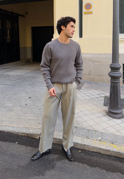Joven con suéter gris acanalado, pantalones beige de pierna ancha y zapatos negros, de pie en la acera de la ciudad cerca de un poste de luz.