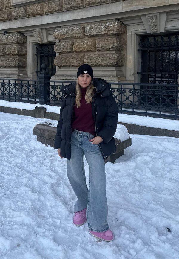 Woman wearing black puffer jacket, burgundy sweater, wide jeans, pink shoes, and black beanie standing on snowy ground in front of stone building.