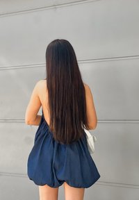 A woman with long dark hair, dressed in a backless navy dress and carrying a white handbag, is standing against a grey wall, facing away.