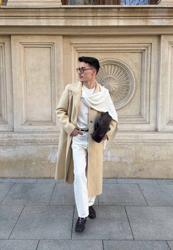 Άνδρας με μπεζ μακρύ παλτό, λευκό πουλόβερ και παντελόνια, με μαύρα παπούτσια και σκούρα γυαλιά ηλίου, περπατάει στο πεζοδρόμιο της πόλης.