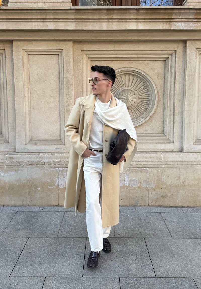 Άνδρας με μπεζ μακρύ παλτό, λευκό πουλόβερ και παντελόνια, με μαύρα παπούτσια και σκούρα γυαλιά ηλίου, περπατάει στο πεζοδρόμιο της πόλης.