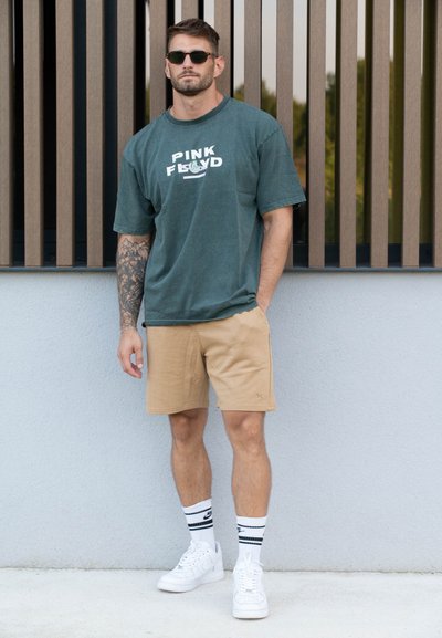 Camiseta verde de gran tamaño con el texto "PINK FLOYD", pantalones cortos beige, zapatillas blancas y calcetines a rayas. Tatuaje distintivo en el brazo izquierdo.