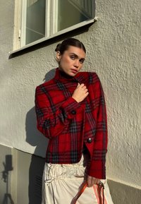 Rød og sort ternet uldblazer med høj krave og to sorte knapper, kombineret med en cremefarvet nederdel og taske med orange detaljer.