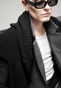 Colletto a maglia intrecciata nera e cappotto in lana grigia esposti con una camicia grigia chiara sotto. Presenta occhiali da sole neri eleganti e piccoli orecchini.