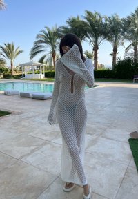 Abito bianco a maniche lunghe con design in rete, scollatura a V profonda e dettagli tagliati. Il modello è a piedi nudi su piastrelle, con una piscina sullo sfondo.