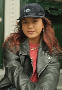 Black corduroy cap featuring a white logo, a glossy-textured leather jacket, layered necklaces, and a red shirt, set against a green background.