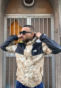 Beige and black waterproof jacket featuring a camouflage pattern, zippered pockets, and the North Face logo on the chest.