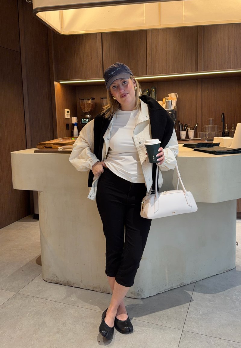 Femme portant une casquette de baseball, une veste blanche, un pantalon noir et des chaussures, appuyée contre un comptoir beige, tenant une tasse de café et un sac blanc dans un café.