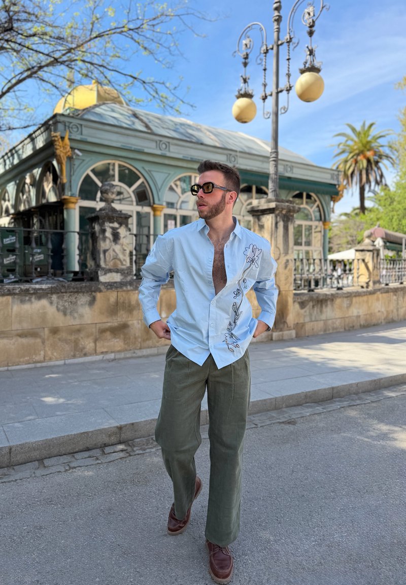 Man wearing sunglasses, light blue shirt with floral design, and olive pants walking on a street with ornate building and lamp posts in background.