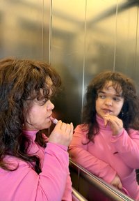 Osoba nanosi sjajilo za usne u dizalu; nosi ružičastu majicu dugih rukava. Zrcalo odražava sjajne metalne zidove i drugi kut.