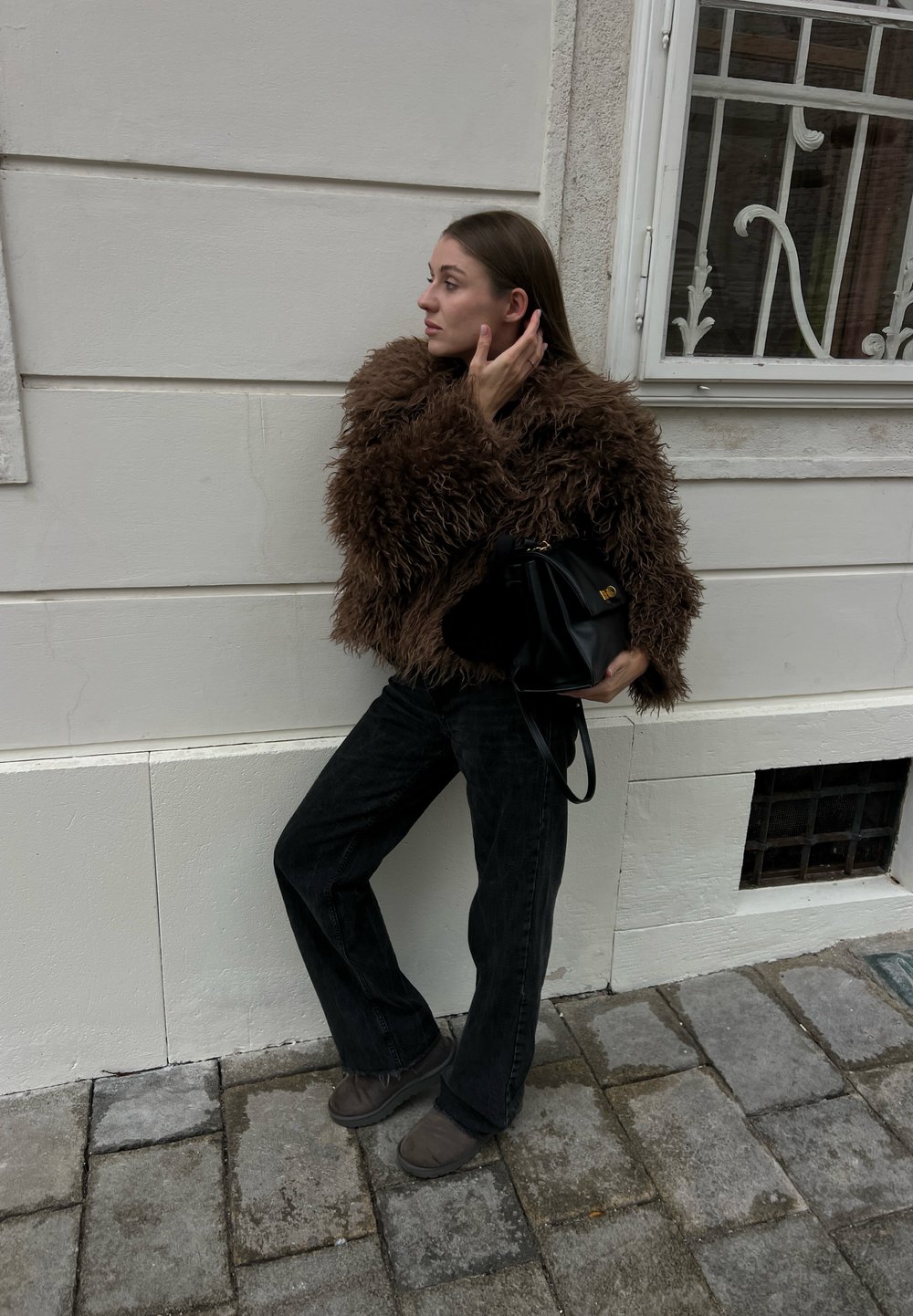 Donna appoggiata a una parete chiara, indossa una giacca marrone pelosa, pantaloni neri e stivali, tiene in mano una borsa nera, si tocca i capelli, in piedi su un pavimento in pietra.