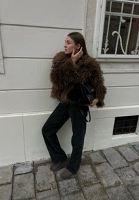 Donna appoggiata a una parete chiara, indossa una giacca marrone pelosa, pantaloni neri e stivali, tiene in mano una borsa nera, si tocca i capelli, in piedi su un pavimento in pietra.