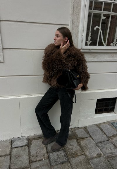 Mujer apoyada contra una pared clara, vistiendo chaqueta marrón de peluche, pantalones negros y botas, sosteniendo un bolso negro, tocándose el cabello, de pie sobre un pavimento de piedra.