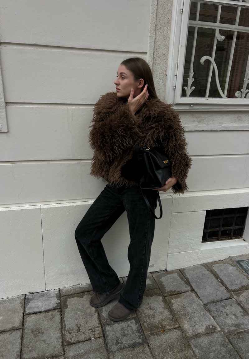 Vrouw leunt tegen een lichte muur, draagt een bruine zachte jas, zwarte broek en laarzen, houdt een zwarte handtas vast, raakt haar aan, staat op stenen bestrating.