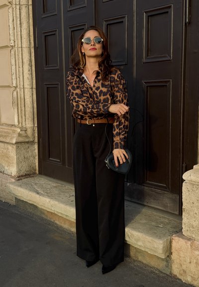 Blusa de estampado de leopardo, pantalones negros de cintura alta, gafas de sol redondas, cinturón marrón y un pequeño bolso negro en forma de corazón. Textura sólida.
