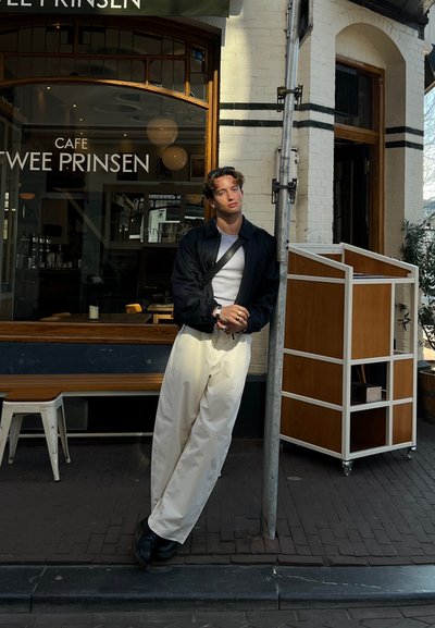 Chaqueta negra sobre una camiseta blanca, pantalones amplios crema, de pie al lado de un poste y una estructura de madera en un entorno de cafetín urbano.