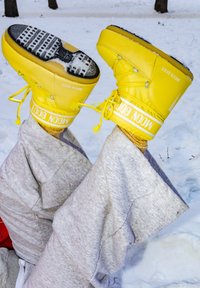 Άτομο ξαπλωμένο στο χιόνι με τα πόδια σηκωμένα, φοράει φωτεινά κίτρινα χειμερινά παπούτσια Moon Boot και γκρι φόρμα.