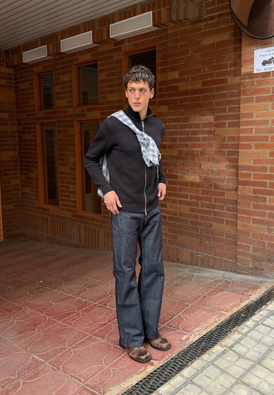 Joven con suéter negro con cremallera, vaqueros de pierna ancha y botas marrones desgastadas, junto a una pared de ladrillo con ventanas y un cartel encima.