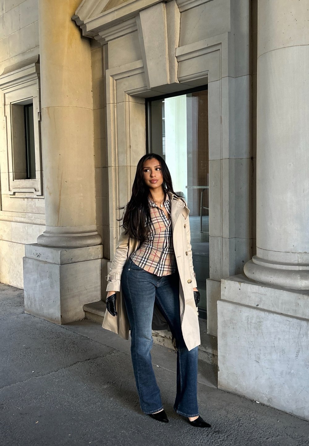 Trench beige sopra una camicia a quadri, abbinata a jeans blu a zampa. Guanti neri e tacchi neri appuntiti completano il look. Colonne in pietra sullo sfondo.