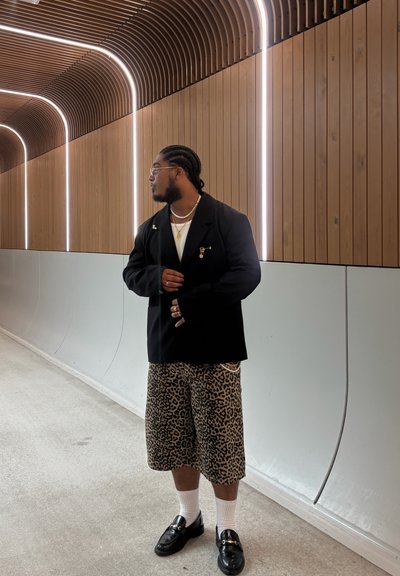 Blazer negro sobre una camisa blanca, combinado con pantalones cortos de leopardo. Mocasines negros con hebillas y calcetines blancos. Accesorios mínimos.
