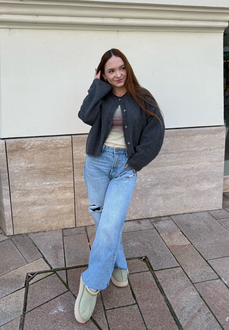 Jeune femme aux longs cheveux bruns portant un cardigan gris foncé, un haut en tie-dye, un jean bleu clair déchiré et des bottes beiges, se tenant sur un pavé carrelé.
