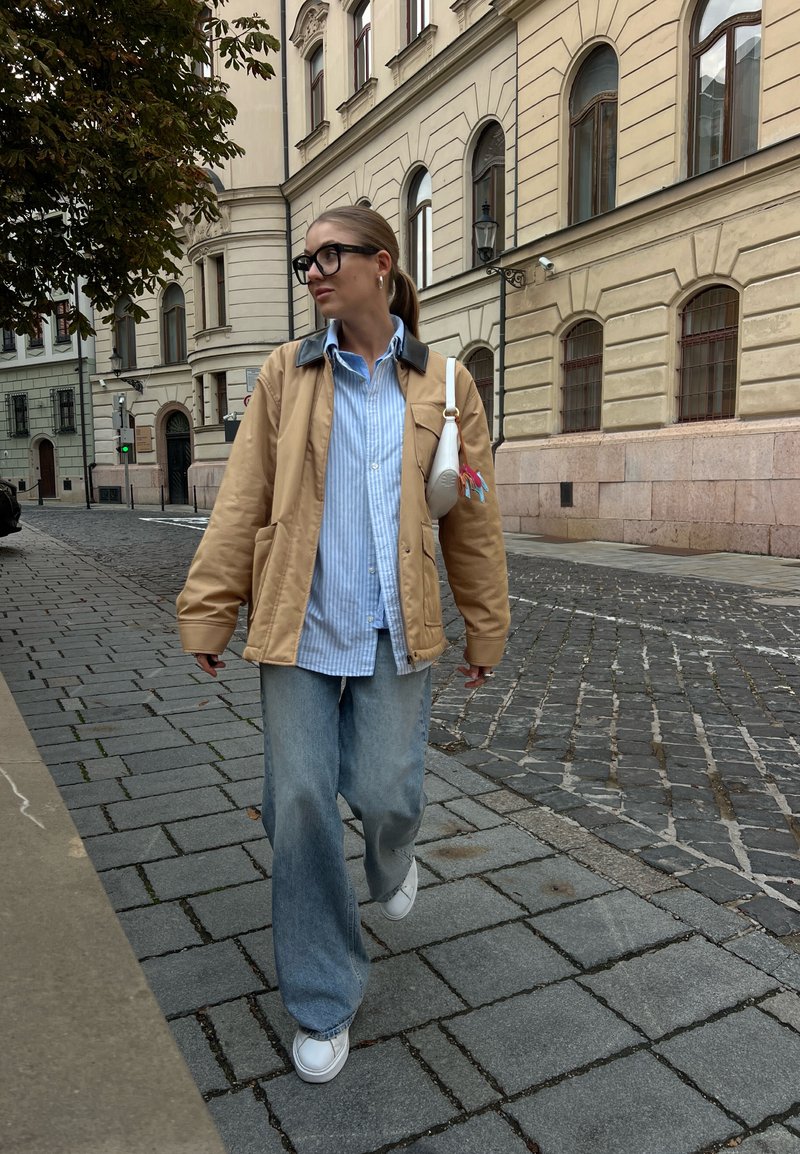 Μπεζ bomber jacket με μαύρο γιακά, ανοιχτό μπλε ριγέ πουκάμισο, φαρδιά ανοιχτόχρωμα τζιν, λευκά αθλητικά παπούτσια και μια μικρή λευκή τσάντα χιαστί.