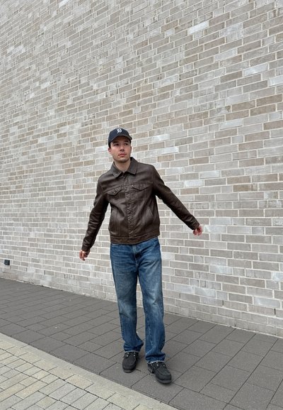 Chaqueta de cuero marrón con cuello, vaqueros azules y zapatos negros, de pie contra una pared de ladrillo de color claro. Estilo casual.