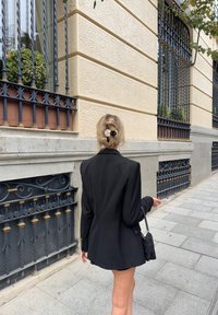 Blazer nero oversize con spalle strutturate e tessuto texturizzato, abbinato a una piccola borsa nera con frange. Capelli chiari raccolti in uno chignon.