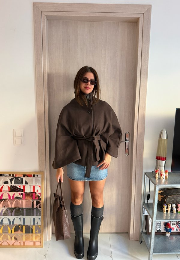Une femme portant des lunettes de soleil, une cape brune ceinturée, une jupe en jean et des bottes hautes jusqu’aux genoux pose près d’une porte, un sac à main à la main.