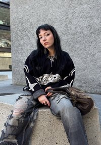 Czarny, oversize'owy sweter z białym wzorem graficznym, poszarpane szare dżinsy z dużymi rozdarciami oraz brązowa torebka z futrzanym akcentem.