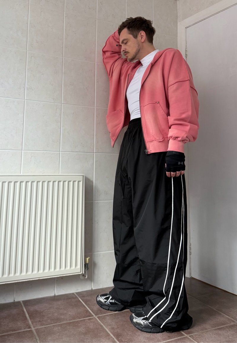 Roze zip-up hoodie met een oversized pasvorm, een wit T-shirt eronder en zwarte baggy broek met witte zijkanten. Zwarte sportieve schoenen met een gestructureerd ontwerp.