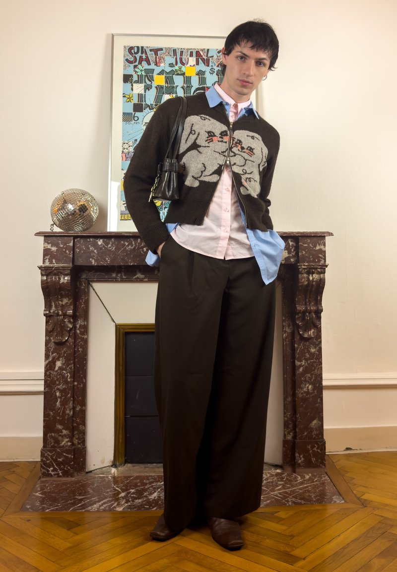 Junge Person trägt eine schwarze Strickjacke mit Tiermotiv, mehrere Schichten von Hemden, weite schwarze Hosen, braune Stiefel und steht vor einem Marmorkamin.