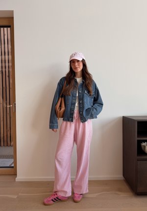 Mujer con gorra rosa claro, chaqueta vaquera, camiseta blanca, pantalones sueltos rosas y zapatillas rosas, de pie en el interior frente a una pared blanca.