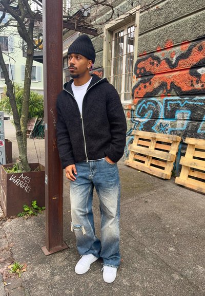 Hombre con gorro negro, chaqueta negra con cremallera, camiseta blanca, vaqueros desgastados con rotos y zapatillas blancas, de pie junto a una pared cubierta de grafitis y palés de madera.