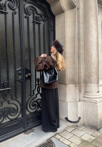 Manteau marron oversize, sac noir et jupe noire, debout près d'une grille en fer noir décorative. Textures remarquables et finitions lisses présentes.