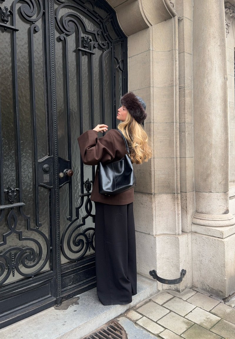 Manteau marron surdimensionné, sac noir et jupe noire, debout près d'une grille décorative en fer noir. Textures notables et finitions lisses présentes.