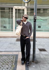 Cardigan grigio sopra una camicia bianca, pantaloni larghi neri, scarpe nere, borsa a tracolla beige, cappello con stampa leopardata, occhiali da sole, in piedi accanto a un palo.