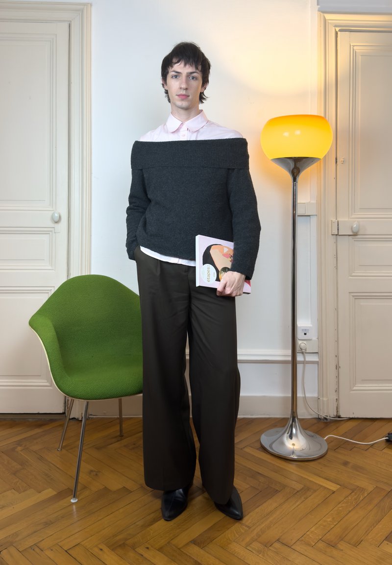 Person wearing dark off-shoulder sweater over white shirt and wide pants, holding a book, standing near green chair and glowing floor lamp.