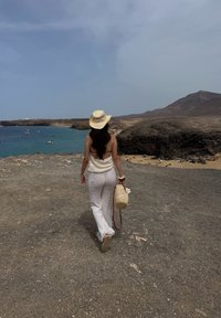 Top croșetat alb, lejer, și pantaloni, pălărie din fân, geantă țesută bej cu floare, îndreptată spre o coastă stâncoasă și mare albastră sub un cer înnorat.