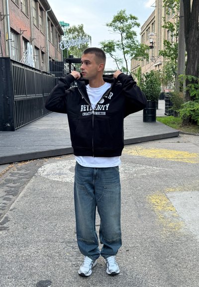 Sudadera con cremallera negra y texto blanco, colocada sobre una camiseta blanca. Jeans de mezclilla azul anchos y zapatillas plateadas. Fondo urbano con vegetación.
