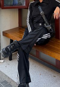 Sort træningsdragt med hvide striber, bestående af en lynlåsjakke og løse bukser, parret med kraftige sorte sneakers. Sidder på en træbænk.