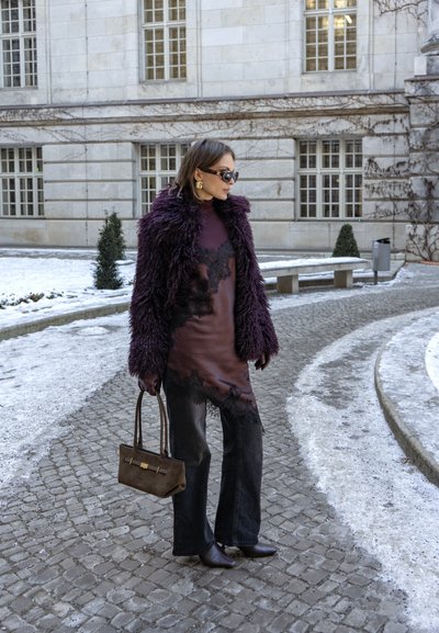 Mujer con abrigo de piel morado, vestido con encaje en los bordes, pantalones negros y gafas de sol, sosteniendo un bolso marrón, de pie en un camino empedrado cubierto de nieve.